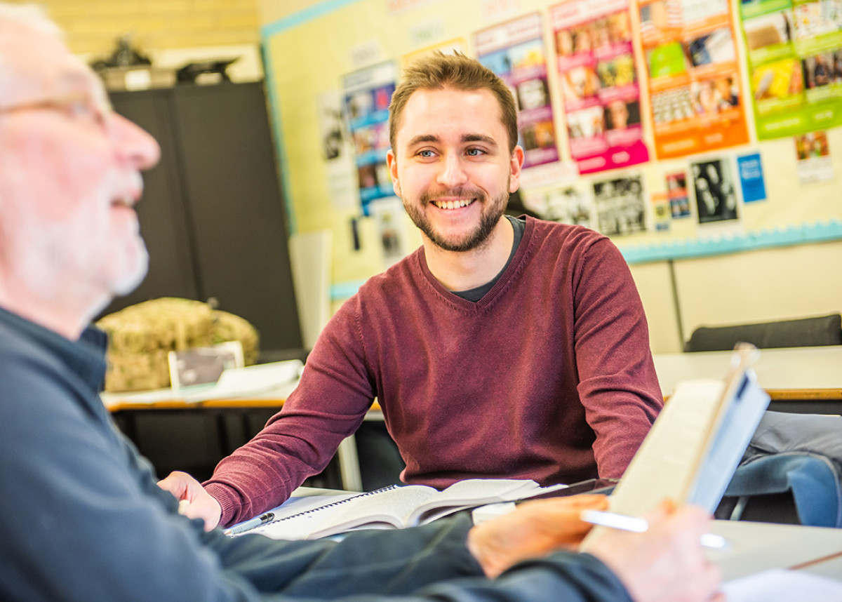 A Higher Education student sat at a table, smiling. Another student is sat close by, book in hand.